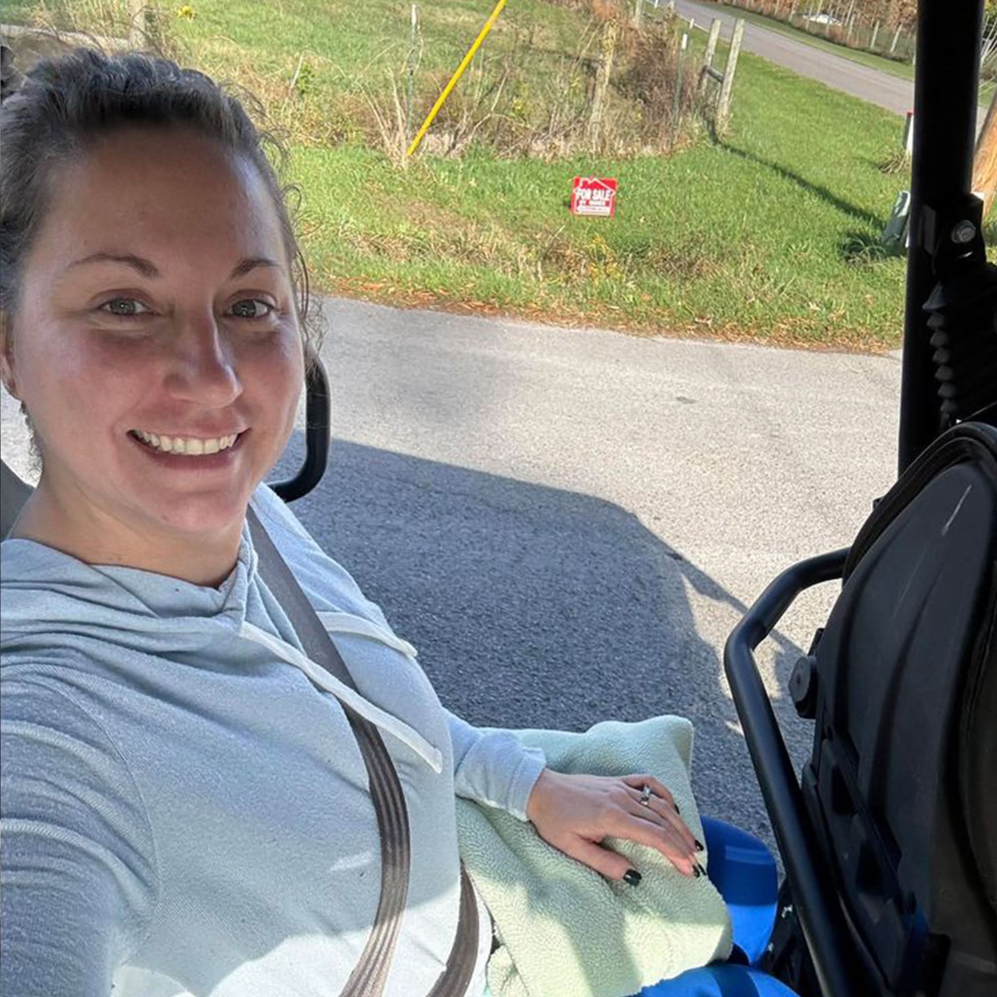Smiling woman in a light hoodie sitting in a vehicle outdoors on a sunny day