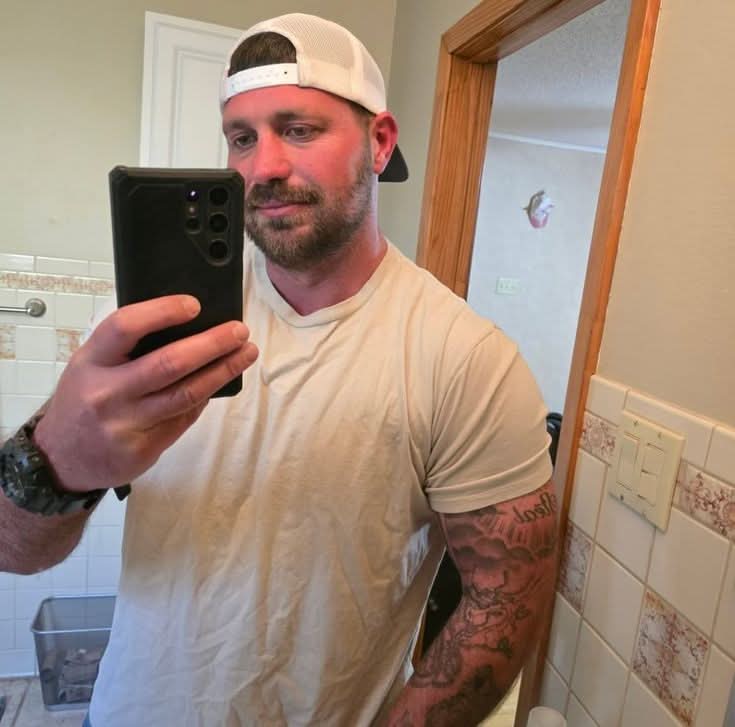 Man in white t-shirt and backwards cap taking bathroom mirror selfie, showing health and fitness